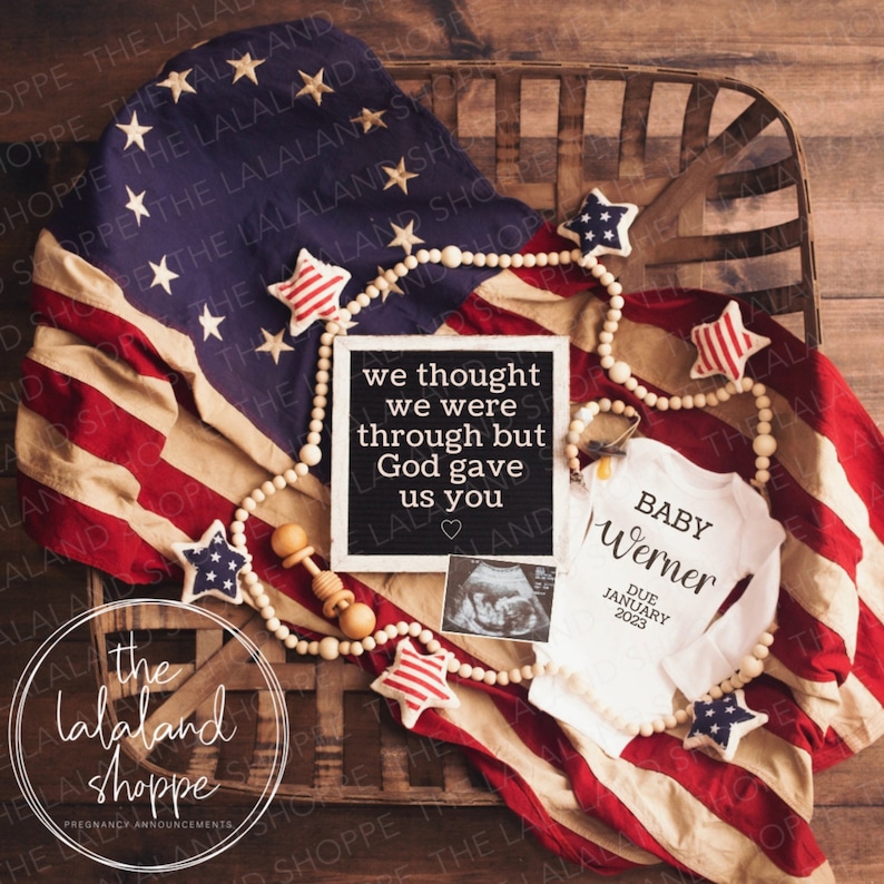 May include: A pregnancy announcement with a white onesie that says "Baby Werner Due January 2023" and a chalkboard sign that says "We thought we were through but God gave us you". The onesie and sign are on a red, white, and blue American flag with a wooden bead garland with red, white, and blue stars.