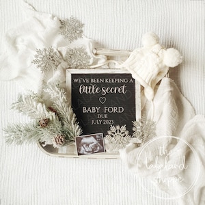 Puede incluir: Un letrero de pizarra con el texto "We've been keeping a little secret" y un corazón. Debajo del texto se lee "Baby Ford Due July 2023". El letrero está rodeado de mantas blancas, un gorro blanco con pompones y copos de nieve plateados.
