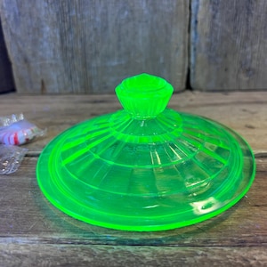 May include: A bright green glass lid with a central knob and a circular, ridged design. The lid is sitting on a wooden surface. Wrapped candies are visible in the background.