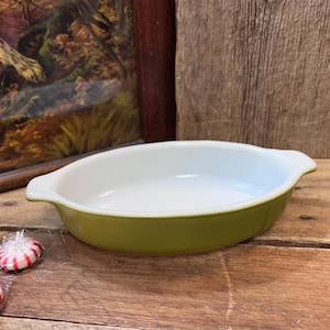 May include: Oval-shaped, vintage baking dish with a white interior and olive green exterior. The dish has two small handles and is likely made of ceramic or glass. A few wrapped candies are in the foreground.