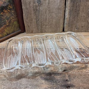 May include: Clear glass corn-shaped serving dish. The dish has six corn cob impressions and a wavy edge. The dish is made of clear glass and is designed to hold corn on the cob. The dish is a vintage item.