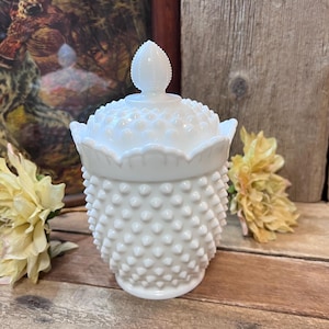 May include: A white hobnail milk glass jar with a lid. The jar has a textured surface with small, rounded bumps. The lid is topped with a decorative finial. The jar is sitting on a wooden surface with yellow flowers in the background.