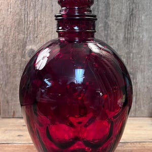 May include: A deep red glass decanter with a bulbous body and a decorative, swirled stopper. The decanter has a rounded shape and a smooth, reflective surface. The stopper is a matching deep red color and has a spiral design. The background is a wooden surface.