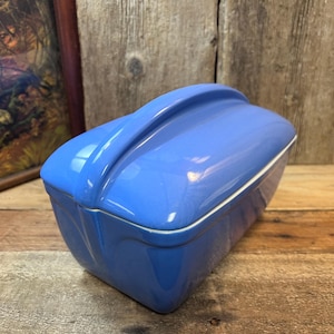 May include: A light blue ceramic butter dish with a curved handle on the lid. The rectangular dish has a white trim and sits on a wooden surface. The background includes a framed picture and wooden planks.