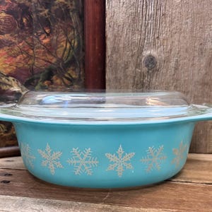 May include: A vintage turquoise Pyrex casserole dish with a clear glass lid. The dish features a snowflake pattern in white. The dish is on a wooden surface, with a painting in the background.