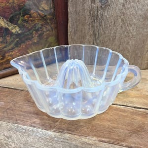 May include: A clear, opalescent glass citrus juicer with a scalloped rim and a built-in reamer. The juicer has a handle and is designed for easy juice extraction. The glass has a slight iridescence, giving it a soft, pearly appearance. A vintage kitchen tool.