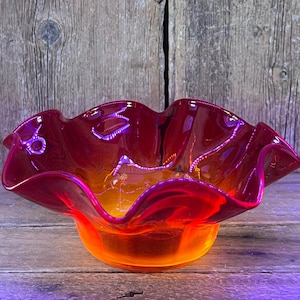 May include: A glass bowl featuring a vibrant gradient color scheme that transitions from a deep red at the ruffled, scalloped edges to a bright orange at the base. The bowl has a smooth, glossy finish and a wide, open design with a curved rim.