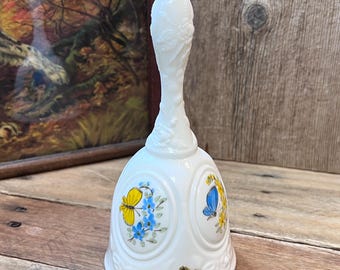 May include: A white milk glass bell featuring a tall, slender handle with embossed floral details. The bell body displays oval panels with hand-painted yellow and blue butterfly motifs and floral accents. A gold Fenton sticker is on the base.