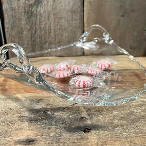 May include: A clear glass serving tray with a wavy edge and two handles. The tray is filled with red and white peppermint candies.
