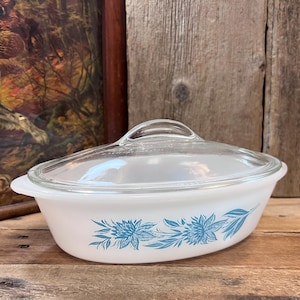 May include: An oval white milk glass casserole dish with a clear glass lid. The exterior of the dish features a blue floral pattern consisting of stylized flowers and leaves. The dish has integrated handles on both sides and a smooth finish.