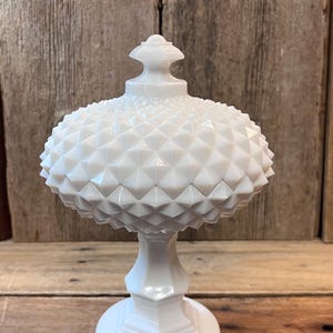 May include: A white, vintage-style candy dish with a textured, diamond-patterned lid. The dish has a pedestal base and a rounded, textured body. The overall design is elegant and classic, suitable for display or serving. The dish is made of opaque white glass.