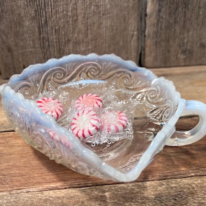 May include: A clear, opalescent glass dish with a handle, filled with wrapped peppermint candies. The dish has a scalloped edge and ornate embossed detailing. The candies are red and white striped.