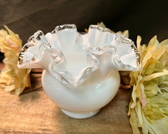 May include: A white, ruffled-edge glass bowl with a gold trim. The bowl has a round base and a flared, wavy rim. The bowl is set against a dark background with dried flowers.