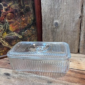 May include: A clear, rectangular glass butter dish with a lid. The dish has a ribbed design on the sides and a small handle on the lid. The dish is sitting on a wooden surface.