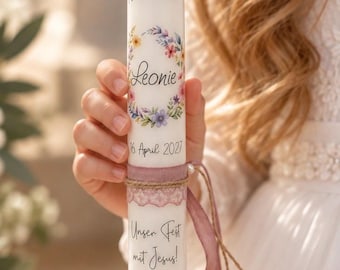 May include: A white cylindrical candle featuring the text 1. Heilige Kommunion, the name Leonie, the date 16. April 2027, and the phrase Unser Fest mit Jesus! It is decorated with a floral wreath, a cross, a mauve fabric band, and a thin ribbon.