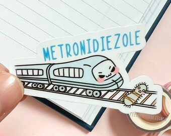 May include: A white sticker with a blue train and the word "METRONIDIZOLE" in blue text. The train has a cartoon face with a frown and blush. A small cartoon bug is on the tracks behind the train.
