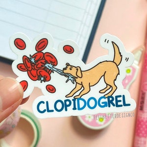 May include: A white sticker with a cartoon illustration of a dog biting a cluster of red blood cells. The text "CLOPIDOGREL" is written in blue letters below the dog. The sticker is held in a hand.