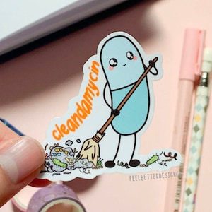 Puede incluir: Un personaje de p&iacute;ldora azul con una cara amigable sosteniendo una escoba y barriendo un mont&oacute;n de g&eacute;rmenes coloridos. El texto "cleandamycin" est&aacute; escrito en naranja en la pegatina. La pegatina est&aacute; sobre una superficie rosa.