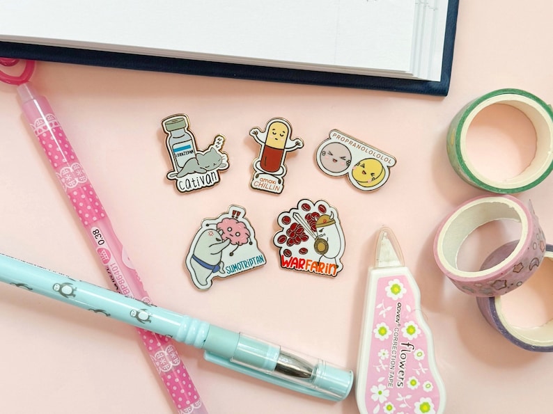 May include: A collection of six enamel pins featuring various medical-themed illustrations. The pins are on a pink surface with a blue and white notebook, a pink pen, and colorful washi tape.