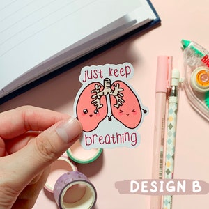 May include: A white sticker with a pink cartoon illustration of two lungs with smiling faces. The text "just keep breathing" is written below the lungs.
