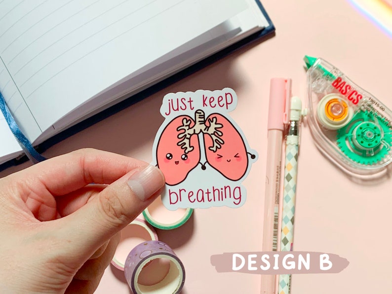 May include: A white sticker with a pink cartoon illustration of two lungs with smiling faces. The text "just keep breathing" is written below the lungs.
