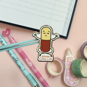 May include: A gold-colored enamel pin featuring a cartoon pill character with a red body and a yellow head. The pill character has a smiling face and is holding its arms up in the air. The text "amoxichiLLIN" is printed below the pill character.