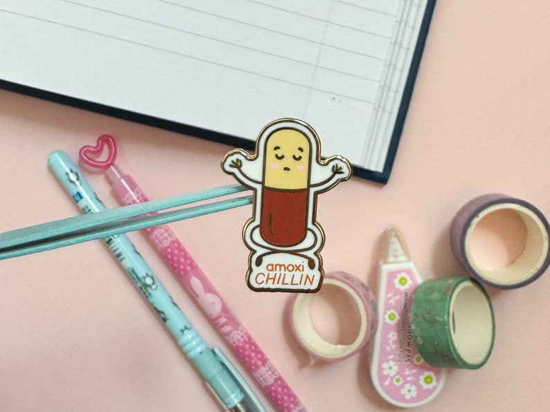 May include: A gold-colored enamel pin featuring a cartoon pill character with a red body and a yellow head. The pill character has a smiling face and is holding its arms up in the air. The text "amoxichiLLIN" is printed below the pill character.