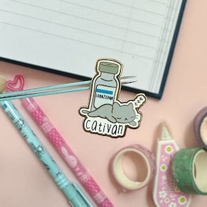 May include: Gold-toned enamel pin featuring a cartoon cat sleeping on a bottle labeled "Lorazepam" with the text "Cativan" below.