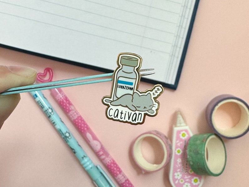 May include: Gold-toned enamel pin featuring a cartoon cat sleeping on a bottle labeled "Lorazepam" with the text "Cativan" below.