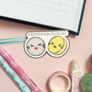 May include: A gold-colored enamel pin with the text "PROPRANOLOLOLOL" in pink and white letters. The pin features two cartoon faces, one pink and one yellow, with smiling expressions and closed eyes. The faces are connected by a blue and white striped bar.