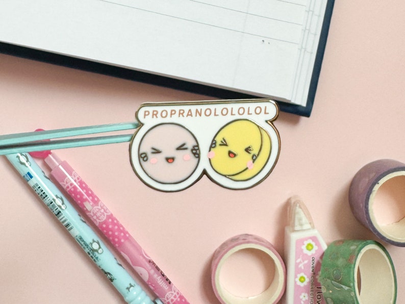May include: A gold-colored enamel pin with the text "PROPRANOLOLOLOL" in pink and white letters. The pin features two cartoon faces, one pink and one yellow, with smiling expressions and closed eyes. The faces are connected by a blue and white striped bar.
