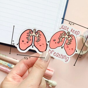 May include: Two pink cartoon lungs with smiling faces and the text "just keep breathing" on a white background. The sticker is 5.5 cm wide and 5.2 cm tall.