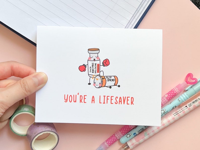 May include: A greeting card with a cartoon illustration of a vial of naloxone knocking out a pill labeled "RIP Opioid". The card reads "You're a Lifesaver".