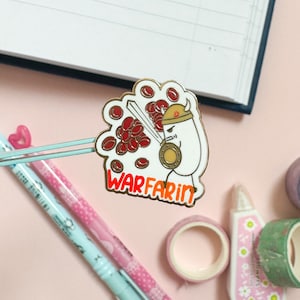 May include: A gold-colored enamel pin with a cartoon illustration of a white character wearing a helmet and holding a sword. The character is surrounded by red blood cells. The text "WARFARIN" is written in orange below the character.