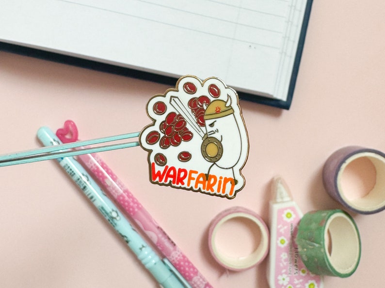 May include: A gold-colored enamel pin with a cartoon illustration of a white character wearing a helmet and holding a sword. The character is surrounded by red blood cells. The text "WARFARIN" is written in orange below the character.