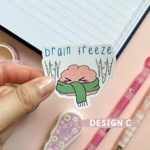 May include: A sticker with a cartoon brain wearing a green scarf and surrounded by icicles. The text "brain freeze" is above the brain. The sticker is labeled "DESIGN C".