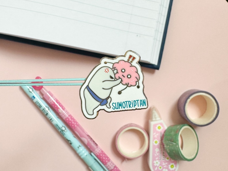 May include: A white enamel pin with a cartoon illustration of a white creature with a blue bow tie and a pink brain with a surprised expression. The text "SUMOTRIPTAN" is below the illustration.
