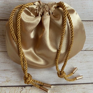 May include: A champagne-colored satin drawstring pouch featuring a smooth, lustrous finish. The bag is cinched at the top with two thick, twisted gold-toned rope cords that hang down, each finished with a polished metallic cylindrical end cap.