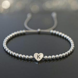 May include: Silver beaded bracelet with a heart-shaped charm. The charm features the letter "K" in the centre. The bracelet has an adjustable sliding knot closure. The beads and charm are silver in colour.