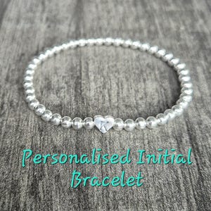 May include: A silver beaded bracelet with a small heart charm. The charm has a single initial engraved on it. The bracelet is on a wooden surface. "Personalised Initial Bracelet" is written in teal text below the bracelet.