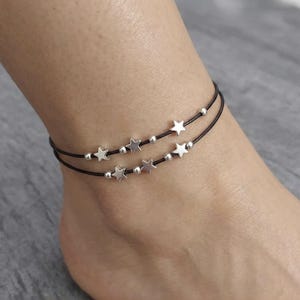 May include: Ankle bracelet featuring two strands of black cord adorned with silver-colored star charms and small beads. The bracelet is designed to be worn around the ankle. The stars are evenly spaced along the cord.