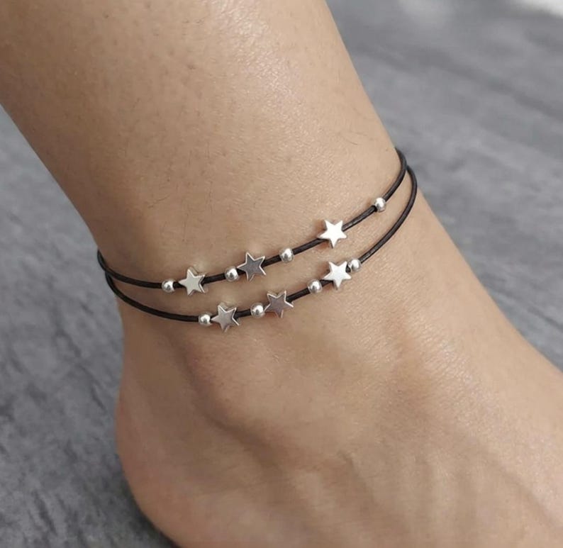 May include: Ankle bracelet featuring two strands of black cord adorned with silver-colored star charms and small beads. The bracelet is designed to be worn around the ankle. The stars are evenly spaced along the cord.
