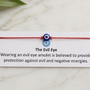 May include: A red string bracelet featuring a blue and white evil eye charm. The bracelet is presented on a white card with the text "The Evil Eye" and a description about protection from negative energies.