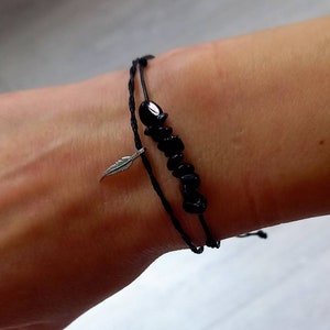 May include: A black cord bracelet with a silver feather charm and black beads. The bracelet is tied around a person's wrist.
