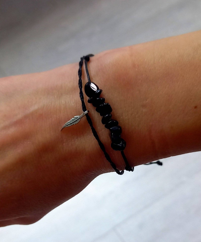May include: A black cord bracelet with a silver feather charm and black beads. The bracelet is tied around a person's wrist.