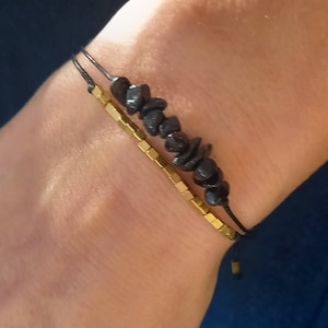 May include: Two bracelets on a wrist. The top bracelet is made of black beads and a black cord. The bottom bracelet is made of gold square beads and a black cord.