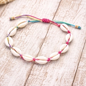 May include: A colourful bracelet made with white cowrie shells and a rainbow-coloured cord. The shells are linked with pink thread. The cord is adjustable. The bracelet has a bohemian style and evokes a beach theme.