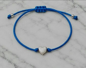 Könnte beinhalten: Ein blaues Kordelarmband mit einem mittigen herzförmigen Anhänger in Silberoptik. Das Armband verfügt über einen verstellbaren Schiebeknotenverschluss und zwei kleine silberfarbene Perlen an den Enden der Kordel für einen flexiblen Sitz.
