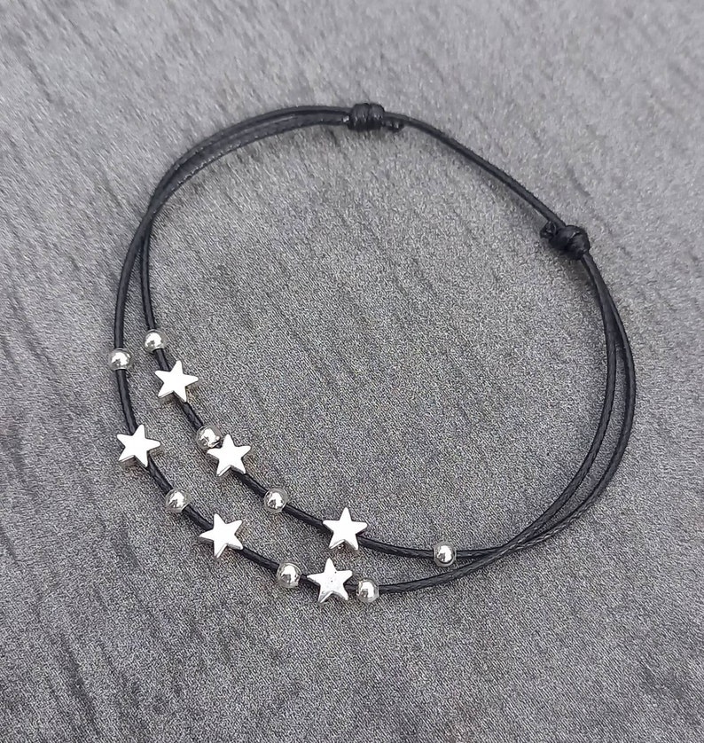 May include: A black cord bracelet featuring two strands adorned with silver-colored star charms and small beads. The bracelet is knotted at the ends, creating a closed loop. The jewelry is displayed on a gray surface.