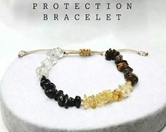 Può includere: Un bracciale con segmenti di pietre scheggiate trasparenti, nere, gialle e marroni su un cordino beige con nodo scorsoio regolabile. Il testo PROTECTION BRACELET è stampato sopra il gioiello con un carattere nero.